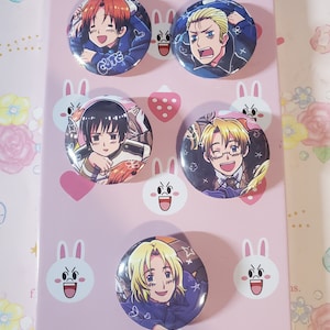 May include: A collection of five round anime character button pins. Each pin features a different character with varying hair colors and expressions. The pins are arranged on a pink surface decorated with bunny and strawberry designs.