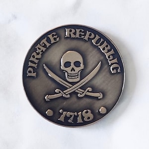 May include: A round, bronze-colored coin with the words "Pirate Republic" around the edge. In the center is a skull and crossbones with the year "1718" below.