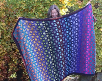 Op de afbeelding: Een kleurrijk gehaakt deken met een geometrisch patroon in tinten blauw, groen, oranje en rood. Het deken wordt vastgehouden door een persoon die voor een groene achtergrond staat.