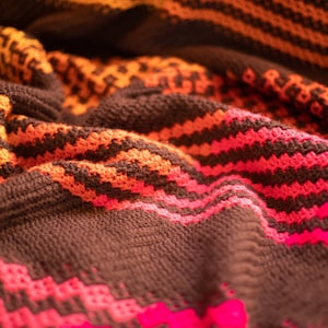 Op de afbeelding: Een close-up van een gestreept gebreid deken in bruin, oranje en roze. Het deken is gevouwen en heeft een zachte, textuur.