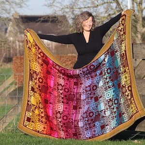 Könnte beinhalten: Eine bunte gestrickte Decke mit einem geometrischen Muster in Rot-, Orange-, Gelb-, Blau- und Grüntönen. Die Decke wird von einer Person in einem schwarzen Shirt gehalten, die vor einem Holzzaun steht.