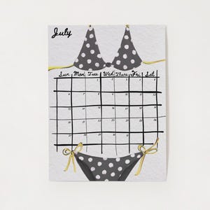 Pode incluir: Ilustração de calendário de julho com um design de biquíni. A parte superior e inferior do biquíni são cinzentas com bolinhas brancas. A grelha do calendário é preta e os dias da semana estão listados no topo. A palavra "Julho" está escrita em itálico.