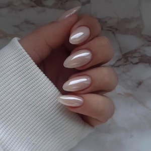 May include: A hand with long, almond-shaped nails painted with a pearlescent, light beige polish. The nails reflect light, creating a shimmering effect. The person is wearing a white sweater.