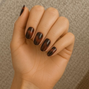 May include: A hand showing oval-shaped nails painted with dark brown and amber nail polish. The nails have a tortoiseshell pattern and a glossy finish. The background is a neutral, textured surface.