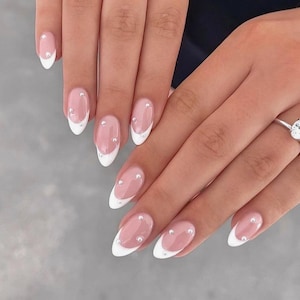 Può includere: Primo piano di una mano con unghie ovali con un design di manicure alla francese. Le unghie hanno una punta bianca, una base rosa trasparente e piccoli abbellimenti argentati. Un anello d'argento con una grande pietra trasparente è sull'anulare.