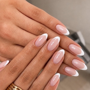 Może przedstawiać: Owalne paznokcie z klasycznym francuskim manicure, z półprzezroczystą, jasnoróżową bazą i wyraźnymi białymi końcówkami. Na palcu serdecznym znajduje się szeroki, polerowany złoty pierścionek o wielowarstwowej, żebrowanej fakturze.
