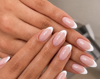 May include: Oval-shaped fingernails featuring a classic French manicure with a sheer, light pink base and crisp white tips. A wide, polished gold ring with a multi-layered, ribbed texture is worn on the ring finger, adding a metallic accent.
