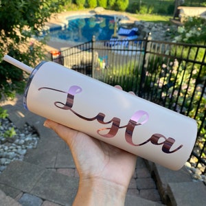 Pode incluir: Um copo de a&ccedil;o inoxid&aacute;vel cor de rosa claro com um design de vinil em ouro rosa que diz "Lyla".