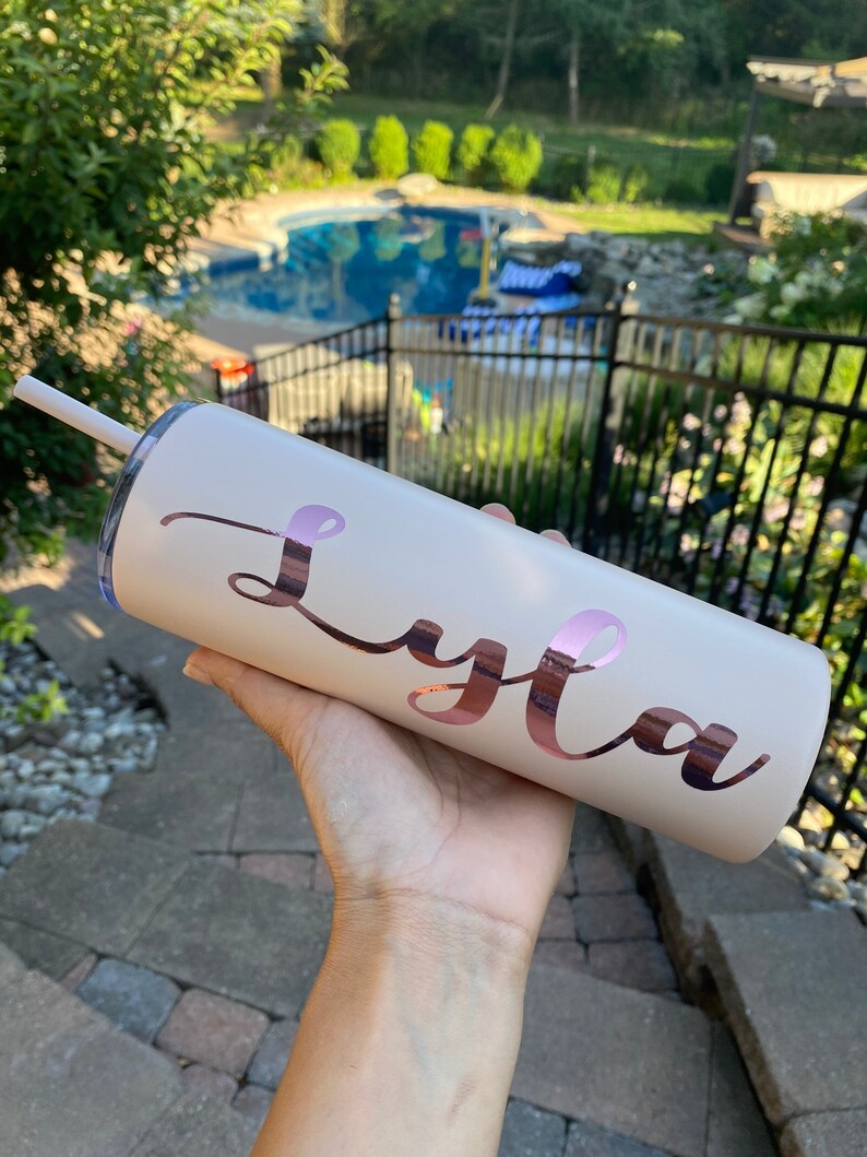 Pode incluir: Um copo de a&ccedil;o inoxid&aacute;vel cor de rosa claro com um design de vinil em ouro rosa que diz "Lyla".
