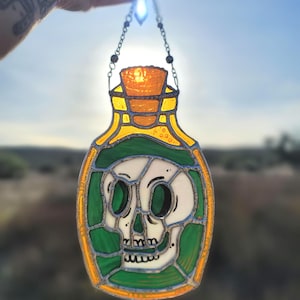 May include: Stained glass potion bottle with a skull design. The bottle features a yellow and green border, a cork, and a chain for hanging. The skull is white with black eye sockets and teeth. The background is a bright, sunny sky.
