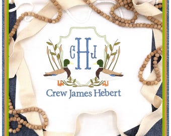 Op de afbeelding: Witte babybody met een blauw en groen geborduurd ontwerp. Het ontwerp toont twee eenden, riet en een monogram met de letters "CHJ". De tekst "Crew James Hebert" is eronder geborduurd. De body is omgeven door houten kralen en crèmekleurig lint.