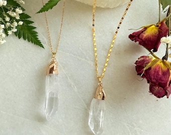 Puede incluir: Dos collares de oro con colgantes de cristal de cuarzo transparente. Los collares están sobre una superficie blanca con hojas verdes y rosas secas.