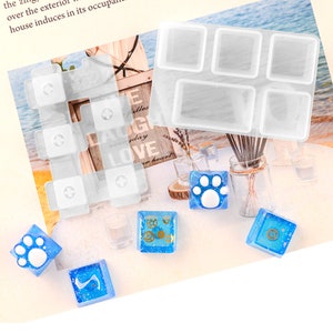 May include: A clear silicone mold with six square and two rectangular cavities. The mold is used to create resin crafts. There are several small blue resin crafts with white paw prints and gold gears on a white surface.