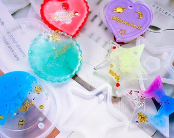 May include: A collection of colorful resin art pieces and molds. Includes heart, star, and circular shapes with zodiac signs like "Sagittarius" and "Aquarius" in gold lettering. The pieces feature glitter and various colors like red, blue, and green.