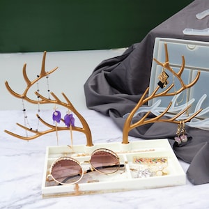 May include: A gold deer antler jewellery organiser with a white marble tray. The tray has compartments for rings, earrings, and sunglasses. The antler stand has multiple branches for hanging necklaces and earrings.