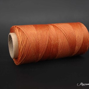 Può includere: Un rocchetto di cordoncino da macramè arancione, avvolto strettamente su un tubo di cartone. Il cordoncino è in fibra naturale, con una superficie leggermente ruvida.