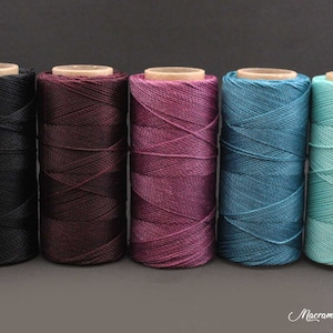 Op de afbeelding: Vijf spoelen macramékoord in verschillende kleuren: zwart, bordeauxrood, paars, teal en mintgroen. De spoelen zijn in een rij op een grijze achtergrond gerangschikt.