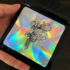 K&ouml;nnte beinhalten: Ein silberfarbener Schl&uuml;sselanh&auml;nger mit einem Kruzifix, einer Medaille des Heiligen Antonius und einer weiteren religi&ouml;sen Medaille. Der Schl&uuml;sselanh&auml;nger wird in einer schwarzen Plastikt&uuml;te mit holografischem Einsatz pr&auml;sentiert.