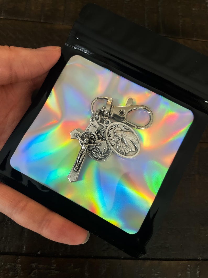 K&ouml;nnte beinhalten: Ein silberfarbener Schl&uuml;sselanh&auml;nger mit einem Kruzifix, einer Medaille des Heiligen Antonius und einer weiteren religi&ouml;sen Medaille. Der Schl&uuml;sselanh&auml;nger wird in einer schwarzen Plastikt&uuml;te mit holografischem Einsatz pr&auml;sentiert.