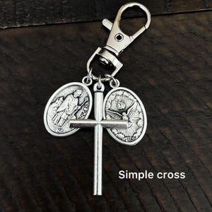 K&ouml;nnte beinhalten: Silberfarbener Schl&uuml;sselanh&auml;nger mit einem Kreuz und zwei ovalen Anh&auml;ngern mit religi&ouml;sen Bildern. Der Schl&uuml;sselanh&auml;nger hat einen Karabinerhaken zum einfachen Befestigen. Der Text "Simple cross" ist auf dem Bild zu sehen.