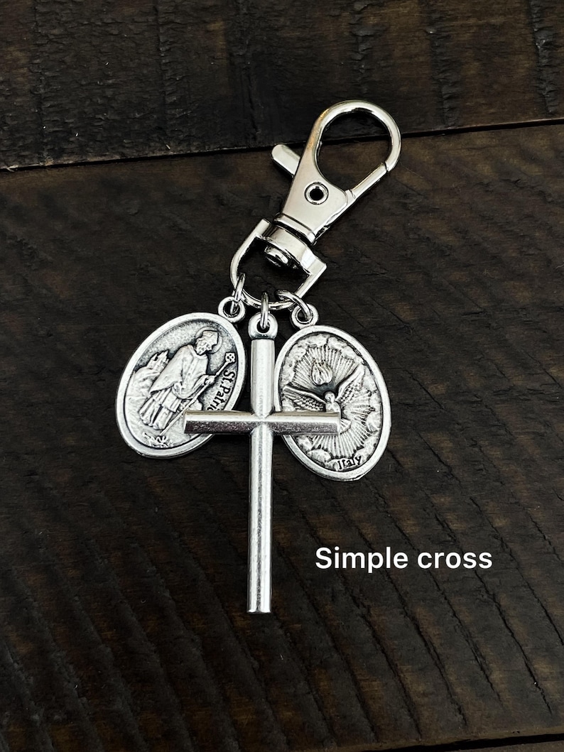 K&ouml;nnte beinhalten: Silberfarbener Schl&uuml;sselanh&auml;nger mit einem Kreuz und zwei ovalen Anh&auml;ngern mit religi&ouml;sen Bildern. Der Schl&uuml;sselanh&auml;nger hat einen Karabinerhaken zum einfachen Befestigen. Der Text "Simple cross" ist auf dem Bild zu sehen.