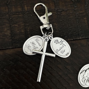 K&ouml;nnte beinhalten: Silberfarbener Schl&uuml;sselanh&auml;nger mit einem Kreuz und zwei ovalen Anh&auml;ngern. Einer der Anh&auml;nger tr&auml;gt die Aufschrift "Come Holy Spirit", der andere eine Rose und die Aufschrift "Protect Us Italy". Der Schl&uuml;sselanh&auml;nger hat einen Karabinerverschluss.