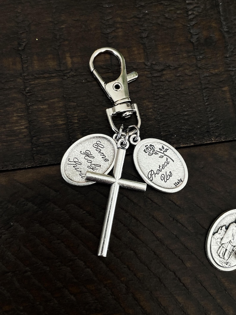 K&ouml;nnte beinhalten: Silberfarbener Schl&uuml;sselanh&auml;nger mit einem Kreuz und zwei ovalen Anh&auml;ngern. Einer der Anh&auml;nger tr&auml;gt die Aufschrift "Come Holy Spirit", der andere eine Rose und die Aufschrift "Protect Us Italy". Der Schl&uuml;sselanh&auml;nger hat einen Karabinerverschluss.