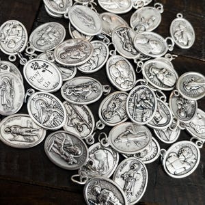 K&ouml;nnte beinhalten: Eine Ansammlung silberfarbener religi&ouml;ser Medaillen mit Darstellungen verschiedener Heiliger und Inschriften. Die ovalen Anh&auml;nger weisen detaillierte Gravuren und kleine Schlaufen zum Anbringen an einer Kette oder einem Armband auf. Die Medaillen liegen auf einer dunklen Holzoberfl&auml;che.