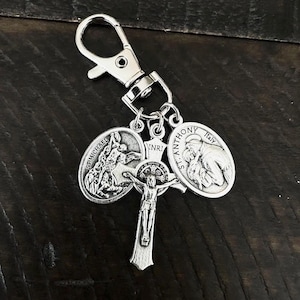 Pode incluir: Chaveiro religioso prateado com um crucifixo e dois medalhões. Um mostra São Miguel, o outro Santo António. O chaveiro inclui um mosquetão para fixação. O crucifixo tem as letras INRI.