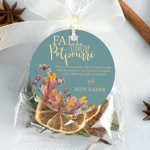 以下が含まれることがあります： 乾燥果物、スパイス、ハーブが入った透明なビニール袋。青い背景に金色の文字で「Fall Stovetop Potpourri」と書かれた丸いタグ。タグにはポプリの使い方の説明が書かれており、「Judy Hanes」の署名があります。