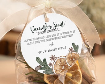 Puede incluir: Una etiqueta redonda blanca con el texto "December Scent Potpourri Simmering Kit" y un dibujo de cítricos secos, palitos de canela y vegetación. La etiqueta está atada a una bolsa de plástico transparente llena de cítricos secos, palitos de canela y vegetación. El texto en la etiqueta dice: "Llene una cacerola pequeña con 2-3 tazas de agua, agregue la mezcla de potpourri y una rodaja de naranja, cocine a fuego lento y rellene el agua según sea necesario. De: TU NOMBRE AQUÍ"