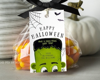 Op de afbeelding: Een wit label met zwarte tekst die "Happy Halloween" en "Have a monstrous good time!" zegt, met een groene Frankenstein-monsterillustratie. Het label is vastgemaakt aan een doorzichtige plastic zak gevuld met candy corn.