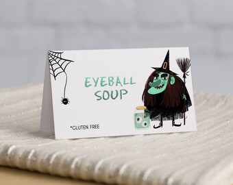 May include: A white card with a cartoon witch holding a jar of eyeballs. The text on the card reads "Eyeball Soup" and "Gluten Free".