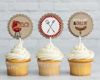 May include: Three cupcakes with white frosting topped with round, brown, glitter-covered paper cupcake toppers. The toppers feature images related to grilling: a red grill with the word "BBQ", crossed grilling tools, and a hot dog with the text "Grillin' & Chillin'".