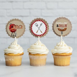May include: Three cupcakes with white frosting topped with round, brown, glitter-covered paper cupcake toppers. The toppers feature images related to grilling: a red grill with the word "BBQ", crossed grilling tools, and a hot dog with the text "Grillin' & Chillin'".