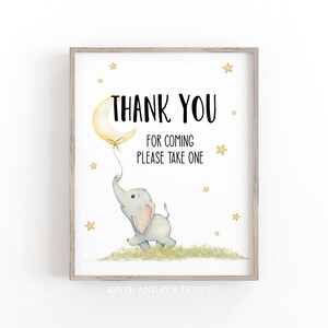 May include: A framed print with a watercolor illustration of a baby elephant holding a moon-shaped balloon. The text reads "THANK YOU FOR COMING PLEASE TAKE ONE" with gold stars scattered around the image. The print is in a light wood frame.