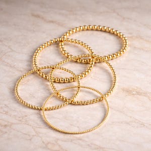 May include: Several gold beaded bracelets of varying sizes are arranged on a marble surface. The bracelets have a shiny, metallic finish and are made of small, round beads. The bracelets are circular and appear to be elastic.