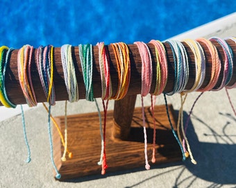 May include: A display of colorful braided bracelets, each tied in a knot at the end, hanging from a wooden stand. The bracelets are arranged in a rainbow pattern.