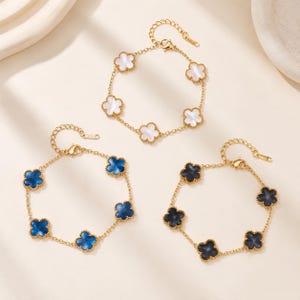 May include: Three gold-tone chain bracelets, each featuring five clover-shaped charms. One bracelet has white iridescent charms, one has deep blue charms, and one has black charms. Each bracelet includes a lobster claw clasp and an extender chain.