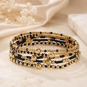 May include: A multi-strand bracelet featuring alternating small gold-toned and black beads, accented with larger, smooth gold-toned spherical beads distributed throughout the design. The bracelet is constructed with a flexible, coiled structure.