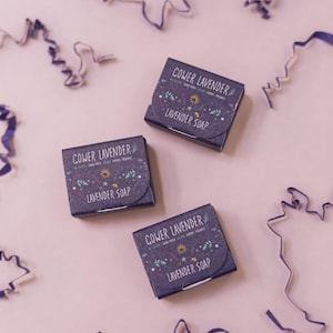 May include: Three boxes of lavender soap are arranged on a pale pink surface, with purple ribbon curls scattered around. Each box is labelled "Cower Lavender" and "Lavender Soap", with floral and sun designs.