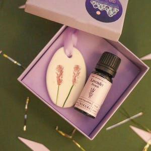 May include: A lavender-themed gift set comprising a ceramic pendant with pink floral designs, a small bottle of French lavender essential oil, and a purple box. The box has a sticker that says "COME LAVENDER".