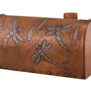 May include: A copper-colored mailbox with a curved top. The front features embossed dragonflies and wheat stalks. The mailbox has a latch and a small, rectangular flag. The overall design is rustic and nature-inspired.