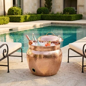 May include: A hammered copper beverage cooler with a lid, filled with ice and bottles of wine and other drinks. The cooler sits on a stone patio next to a pool and lounge chairs. The cooler has a round shape and a handle on the lid.