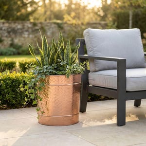 May include: A copper-colored, hammered metal planter filled with green plants and ivy. The cylindrical planter is outdoors, next to a gray cushioned outdoor chair. The background includes a stone wall and green foliage.
