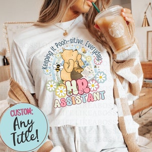 May include: White t-shirt with a Winnie the Pooh illustration and the text "Keeping it Pooh-sitive, Everyday HR Assistant". The shirt has a floral design and a blue and pink custom text box that says "Custom: Any Title!"