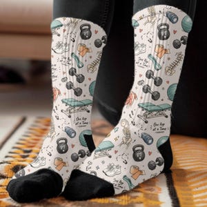 May include: White crew socks featuring a repeating pattern of physical therapy equipment including kettlebells, dumbbells, exercise balls, treatment tables, and anatomical spine illustrations, with the text One Rep at a Time printed in black.