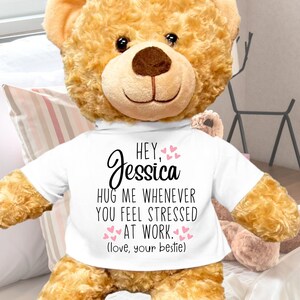 May include: A large, brown teddy bear wearing a white t-shirt with the text "Hey, Jessica hug me whenever you feel stressed at work. (love, your bestie)" printed in black. The bear is sitting on a white and pink striped bedspread. The bear is 22 inches tall.