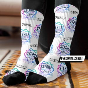May include: White crew socks featuring a repeating pattern of the text 'STERILE PROCESSING CREW' in blue and purple, interspersed with the name 'Sarah' in black. The design includes medical instrument icons and a black toe and heel section.