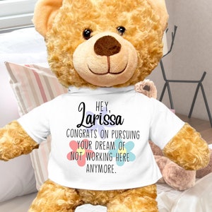 May include: A large, brown teddy bear wearing a white t-shirt with the text "Hey, Larissa Congrats on pursuing your dream of not working here anymore." The bear is sitting on a white bed with a pink and white striped pillow behind it. The bear is 22 inches tall.